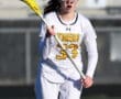 Female lacrosse player in a white jersey with yellow accents, passionately running on the field with a yellow lacrosse stick.