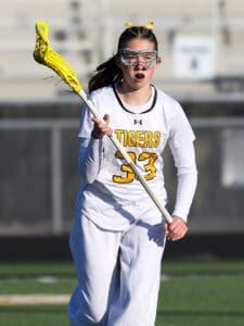 Female lacrosse player in a white jersey with yellow accents, passionately running on the field with a yellow lacrosse stick.