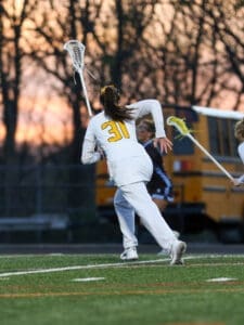 Girl in lacrosse gear running on field at dusk, focusing on receiving a pass with teammates in the background.
