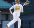Player in a white lacrosse uniform #22 runs, preparing to catch the ball with a lacrosse stick on a sports field.