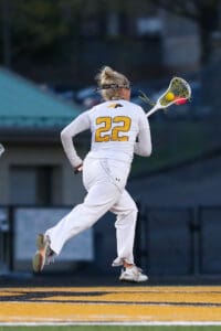 Player in a white lacrosse uniform #22 runs, preparing to catch the ball with a lacrosse stick on a sports field.