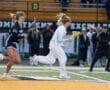 Lacrosse players sprint during a match, showcasing athleticism on the field with teammates and spectators in the background.
