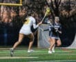 Two female lacrosse players compete on a field at dusk, showcasing athleticism and teamwork during an intense match.