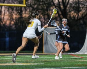 Two female lacrosse players compete on a field at dusk, showcasing athleticism and teamwork during an intense match.