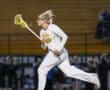 A female lacrosse player in uniform runs with a stick during a night game, showcasing athleticism and focus.