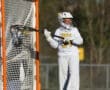 Goalie in action during a lacrosse game, wearing a white uniform and focused on the ball near the goal.