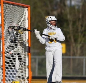 Goalie in action during a lacrosse game, wearing a white uniform and focused on the ball near the goal.