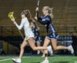 Two female lacrosse players sprinting on the field during an evening game, one shooting the ball toward the goal.