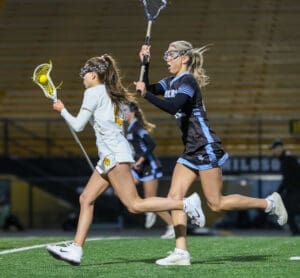 Two female lacrosse players sprinting on the field during an evening game, one shooting the ball toward the goal.