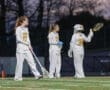 Three female lacrosse players in white uniforms strategizing on a field under twilight, showcasing teamwork and focus.