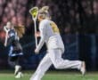 Female lacrosse player in action, wearing white uniform, running on field with stick and ball during a game at dusk.