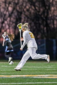 Female lacrosse player in action, wearing white uniform, running on field with stick and ball during a game at dusk.