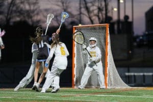 Action shot of a women’s lacrosse game, featuring a player taking a shot on goal while the goalie prepares to defend.