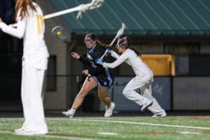 Two female lacrosse players compete for a ball during an evening match, showcasing athleticism and teamwork.