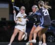 High school girls' lacrosse action shot: players dodging and competing for the ball under stadium lights.