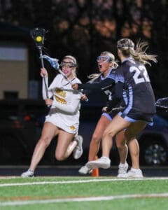 High school girls' lacrosse action shot: players dodging and competing for the ball under stadium lights.