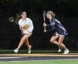 Two female lacrosse players competing fiercely on a night field, one with a stick preparing to pass the ball.