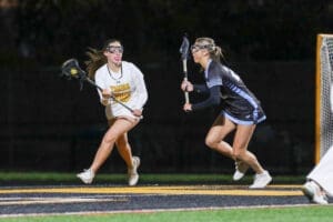 Two female lacrosse players competing fiercely on a night field, one with a stick preparing to pass the ball.
