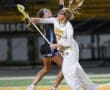 Two female lacrosse players compete for the ball during a game under stadium lights, showcasing athleticism and determination.