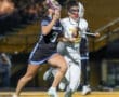Two female lacrosse players in action during a match, one in a blue uniform, the other in white, on a field.