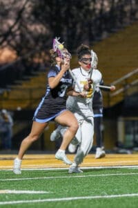 Two female lacrosse players in action during a match, one in a blue uniform, the other in white, on a field.