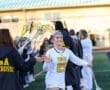 Lacrosse player wearing a white uniform smiles as teammates celebrate her arrival on the field. Team spirit in action!