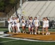 Girls' lacrosse team celebrates on the field, showcasing teamwork and excitement in their white uniforms.