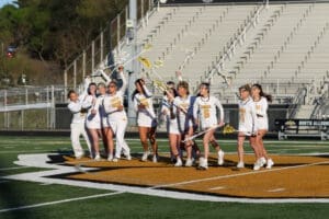 Girls' lacrosse team celebrates on the field, showcasing teamwork and excitement in their white uniforms.