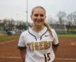 Smiling female softball player in a Tigers jersey stands on the field, showcasing team spirit and athleticism.