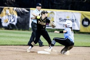 Softball player in black catches a ball to tag a sliding runner in blue. Umpire watches closely during intense game action.