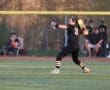 A young female softball player wearing black prepares to throw a ball during a game, with spectators in the background.