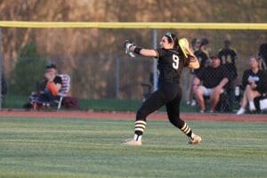 A young female softball player wearing black prepares to throw a ball during a game, with spectators in the background.