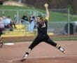 Softball pitcher in action, throwing a pitch on the field with spectators in the background.