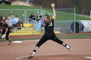 Softball pitcher in action, throwing a pitch on the field with spectators in the background.