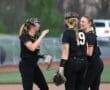 Three softball players in black uniforms engage in a lively discussion on the field during practice.