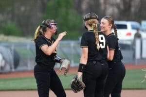 Three softball players in black uniforms engage in a lively discussion on the field during practice.