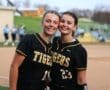 Two softball players in black jerseys with Tigers text smiling together on the field, showcasing team spirit and camaraderie.
