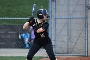A young female softball player in a black jersey and helmet prepares to bat at a game.