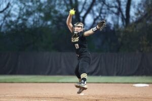 A female softball pitcher in action, wearing a black uniform and preparing to throw a bright yellow ball.
