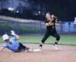 Player sliding into base while another player catches the ball in a competitive softball game under evening lights.