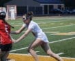 High school girls' lacrosse game action, player in white jersey evading an opponent in red on a turf field.