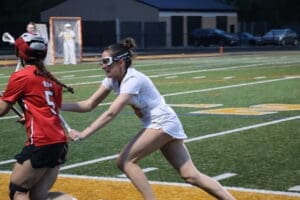 High school girls' lacrosse game action, player in white jersey evading an opponent in red on a turf field.