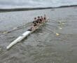 Teenage rowers training on a calm lake, focused on their technique, under a cloudy sky.