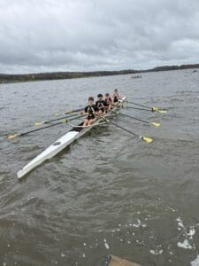 Teenage rowers training on a calm lake, focused on their technique, under a cloudy sky.