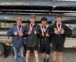 Four rowers proudly display their medals, smiling and giving thumbs up, with boats in the background at a rowing event.
