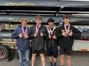 Four rowers proudly display their medals, smiling and giving thumbs up, with boats in the background at a rowing event.