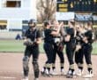 Softball team in black and yellow uniforms gathers on the field for a team meeting, showcasing camaraderie and strategy.