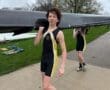 Young male rower carrying a boat, wearing a black and yellow racing suit, with a teammate in the background.