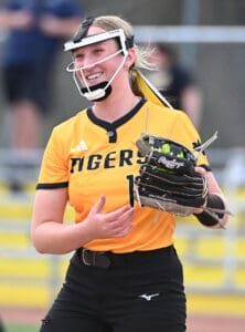 Softball player wearing a yellow Tigers jersey smiles while holding her glove during a game. Team spirit on display!