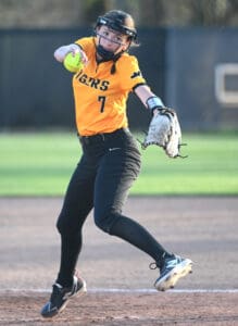 A young female softball player in an orange jersey throws a pitch on a sunny field, showcasing athletic form and focus.
